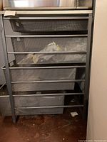Front view of chrome industrial style four-drawer metal storage unit with mesh drawers. Some dirt visible inside drawers and on floor beneath.