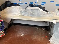 Full view of a rectangular plastic folding table with black folding legs under a white plastic covering, with some clothes or items partially on the table.