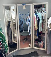 Front view of triple panel mirror showing all three mirrors with two side panels angled inward, hung on a wall with clothing racks reflected in the glass.