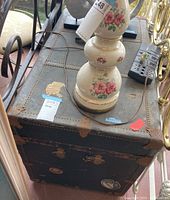 Full view of vintage steamer trunk with floral lamp on top, highlighting black surface with rusted brown metal hardware and corner protectors.