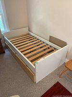 Angled side view of the white laminate twin bed frame showing wooden slats and the drawers partially visible at the bottom.