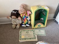 Photo showing two Cabbage Patch Kids dolls together with original yellow box and certificates on carpeted floor near wall.