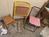 Overview of two metal mesh folding chairs (yellow and brown), two wooden stools, extra wooden stool legs, and a black metal wire magazine rack on a concrete floor in basement.