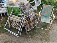 Grouped photo of multiple vintage lawn chairs with metal frames and woven webbing seats in various colors and patterns outdoors on grass and pavement.