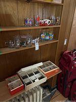 Shelf displaying assorted drinking glasses including Peanuts designs, lobster salt and pepper, and a ceramic mug with lobster design (cracked). Several glasses below in boxes.