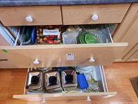 Two kitchen drawers open showing three metal canisters in one with other loose kitchen items, and plastic lids and containers in another drawer