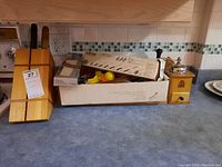 Wide view of wooden knife block with knives, box with utensils and wood cutting board, and manual coffee grinder on countertop.