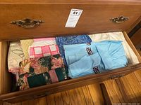 Photo showing opened drawer filled with folded assorted linens including placemats, aprons, and pot holders in varied colors and patterns.