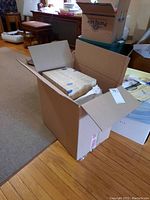Open cardboard box showing folded vintage linens with visible lace and embroidery details, some linens wrapped in decorative paper.