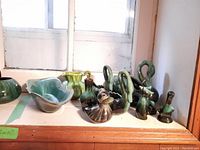 Photo showing various green and brown glazed pottery pieces including swans, cats, duck, pitcher, and plates on a wooden surface by the window.