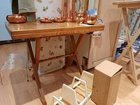 Wooden folding table shown from a front angle with copper items on surface (not included) and floor objects around.