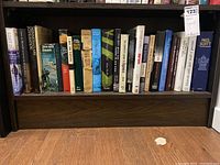 Wide shelf view showing 24 assorted books on a wooden bookshelf. Mix of hardcover and paperback, various colors including blue, white, brown, and black spines.