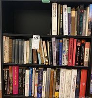 Books arranged on a black shelf, showing front and spine views of many titles including 'William Opper Des Friedens', 'Shadow of Shadows', 'Alec Wiffrand di Calvizio', 'Funny Boy', 'The Classic Book of the New York Dairy 1903-1904', among others.