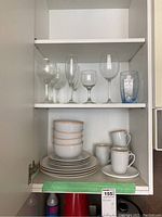 Dishes with gold trim including bowls, plates, cups, missing some saucers, and a blue tumbler glass on a white cabinet shelf