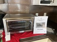 Stainless steel Emeril Lagasse SAFO-001 air fryer and owner's manual placed on a red cloth. Visible glass door and product label.