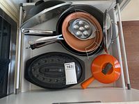 Top-down view of drawer showing orange enamel pot with lid, black cast iron skillet, black oval roaster with lid, copper-colored pan with egg mold, and other pans and utensils.