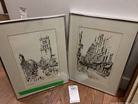 Two framed black and white ink sketches of street scenes leaning against wall