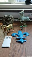 View of all five items on wooden floor: iron horse, brass horse, pewter horse, ceramic owl, blue fleur-de-lis Montreal plaque.