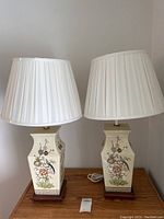 Two lamps with ceramic Chinese style bases and white pleated shades positioned side by side on a wooden surface.