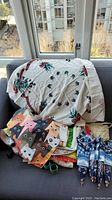 The entire lot of linens piled on a dark couch by a window, showing the tablecloth with floral and tartan print, colorful cat pattern aprons, blue and white patterned napkins, woven placemats, and other linens.