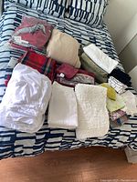 Photo showing folded assorted linens on a bed including a white fitted sheet, bath towels, kitchen towels, face cloths, a red plaid blanket, and a cream bathmat
