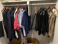View of multiple men's shirts and tops hanging on a closet rod, including dark jackets, light blue button-up shirts, plaid shirts, and darker tops on hangers.