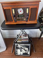 Full front view of the wooden music center showing fabric speaker panels, analog radio dial, control knobs, and the base table it rests on with CDs and vinyl below.