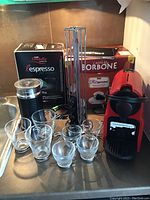 Photo showing red Nespresso coffee maker, two boxes of coffee pods, metal pod holder, black milk frother, and seven clear glass cups on countertop