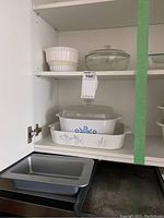 Kitchen cabinet shelf with Pyrex and Corning Ware casserole dishes, white ceramic pieces, and metal baking sheets below