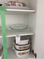 Glass and ceramic serving dishes in white cabinet, including clear Pyrex bowls and Corning Ware casserole dishes with floral patterns.