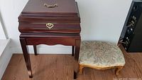 Front view of the wooden storage stand with hinged top and brass handle next to floral upholstered foot rest on hardwood floor.