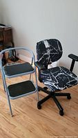 Photo showing both the Herman Miller office chair with black and white marbled removable cover and the two-step metal stool with cushioned black steps and grey metal frame on a wooden floor.