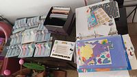Table showing Creative Memories scrapbook supplies including paper ribbons in packaging, decorative paper sheets, sticker sheets, and a binder with cut paper shapes.