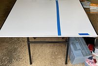 White rectangular drafting table with black metal folding legs, shown flat surface with blue tape strip.