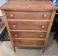 Front view of wooden dresser showing four drawers, round wooden knobs, and caster wheels on legs. Medium brown wood finish with lighter trim around drawer edges.