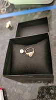 Gold color ladies ring with emerald cut clear stone and separate white pearl on black cushion inside black box.