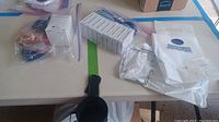 Overview photo showing multiple boxes of scrapbooking adhesives, plastic bag, and black cup holder on table.