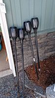 Four black outdoor solar torches with vented heads and stakes leaning against a wall outdoors, showing overall lot contents.