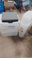 White 3-drawer plastic storage bin on wheels with a gray hard case placed on top, standing on a concrete floor in a garage.
