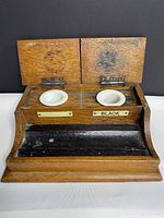 Front view of antique wooden ink well with open lids showing two white porcelain wells and brass labels below them, one labeled 'BLACK'. Dark interior pen rest area.