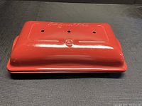 Top and side view of red ceramic baguette baker with ventilation holes and embossed details on lid.