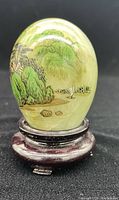 Front view of painted jade egg on wooden stand showing painted landscape with trees and a boat