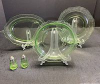 Green depression glass dishes approximately 3 pieces with salt and pepper shakers, displayed on stands with clear lighting showing translucent green glass with Florentine pattern.