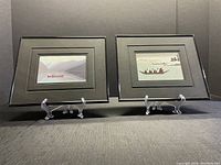 Both framed Inuit art prints displayed side by side on clear plastic stands showing overall condition and framing.