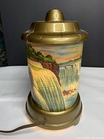 Front view of gold-tone cylindrical lamp with printed Niagara Falls scene illuminated from base.