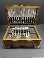Front view of the open wood chest showing dinner knives held vertically, assorted forks and spoons arranged in the lower compartment.