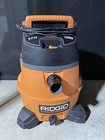 Front view of Ridgid Professional shop vacuum showing orange and black body, power cord wrapped around handle, and hose inlet in front.