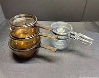 Set of three amber Pyrex Vision cooking pots with lids alongside clear Pyrex double boiler pot with lid on dark surface.