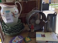 Full lot view showing cabbage plates, ceramic pitcher, metal fan, metal pitcher candlestick holder with red candle, and books on wooden surface.