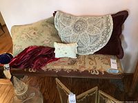 Angled front view of embroidered bench with floral upholstery, decorative nailhead trim, carved dark wood legs, and pillows/blankets resting on top.